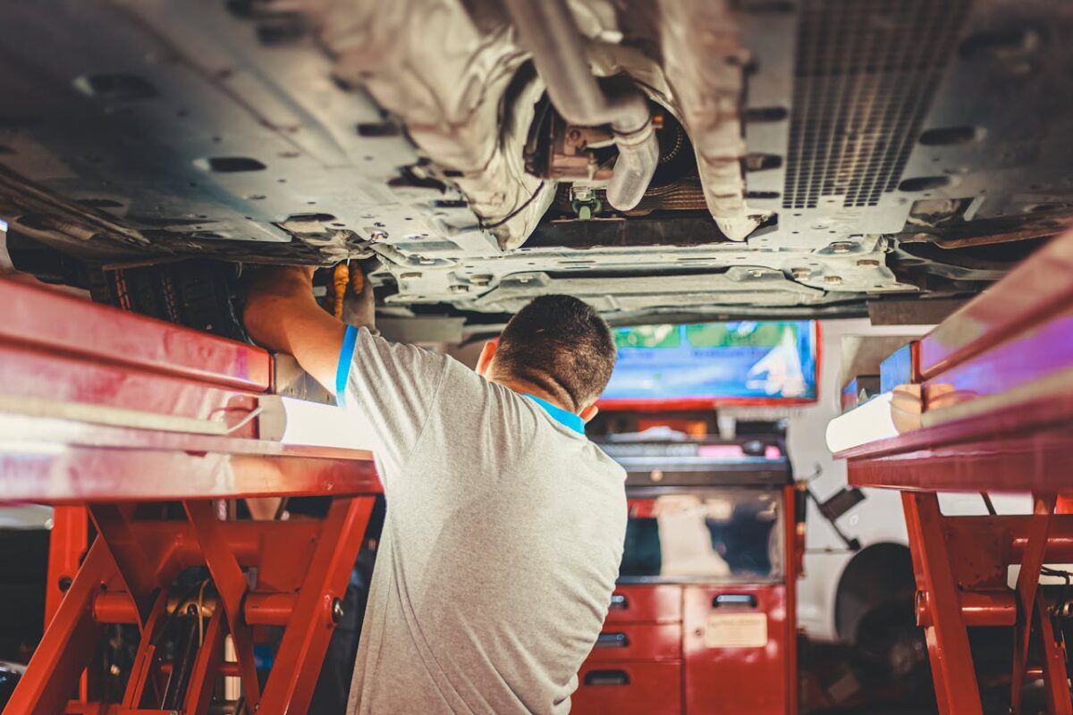 Carrosserie Bras A mechanic works under a lifted car in an auto repair shop, adjusting parts.